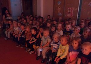Grupa dzieci siedzi na dywanie i ogląda przedstawienie.