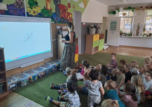 Smyki na dywanie przyglądają się zadaniu wyświetlonym przez Panią Ilonę.