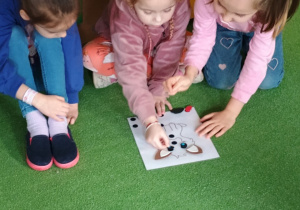 Dziewczynki przyklejają części ciała pieska na kartce.
