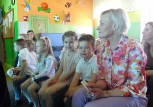 Grupa dzieci i nauczycielka siedzą na krzesełkach.
