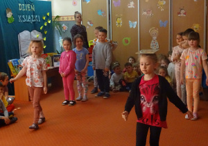 Dzieci w dwóch rzędach idą z książkami na głowie.