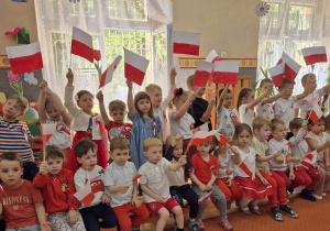 Dzieci siedzą na ławce i trzymają wysoko flagi Polski