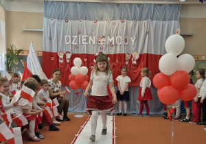 Kolejna dziewczynka z wiankiem na głowie idzie wzdłuż sali.