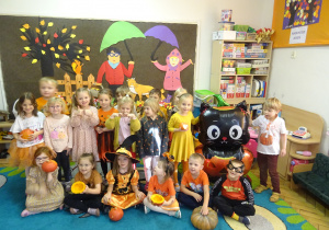 Grupa dzieci siedzi obok siebie i trzyma kolorowe dynie przy sobie.