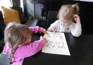 Dwie dziewczynki siedzą przy stoliku z kartkami z wypukłymi kształtami.