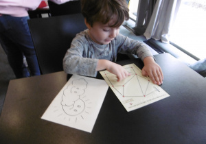 Chłopiec siedzi przy stoliku i trzyma w ręku białe kartki.