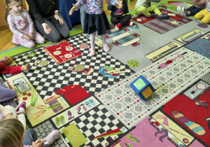 Dziewczynka stoi na dywanie i rzuca kostką z materiału.