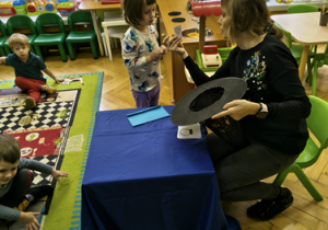 Nauczycielka podaje dziewczynce wróżbę wyjętą z kapelusza.