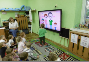 Grupa dzieci siedzi na dywanie przed monitorem z prezentacją multimedialną