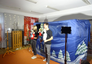 Aktorzy stoją przed dekoracją i trzymają lalki w ręku.