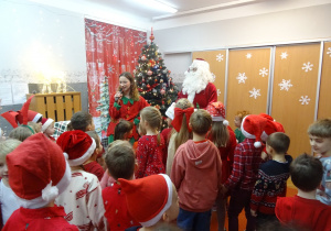 Osoba przebrana za elfa stoi obok Mikołaja i trzyma w ręku mikrofon.