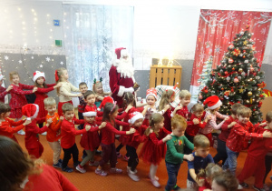 Mikołaj trzyma dzwonek i stoi obok tańczących dzieci.
