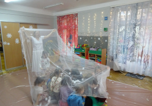 Grupa dzieci siedzi przed białą przezroczystą chustą.