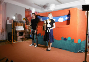 Aktorzy stoją przed dekoracją i trzymają lalki w rękach.