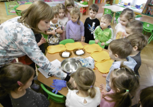 Nauczycielka stoi przy stoliku w otoczeniu dzieci.
