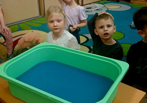 Troje dzieci stoi przy plastikowym pudełku wypełnionym niebieskim płynem.