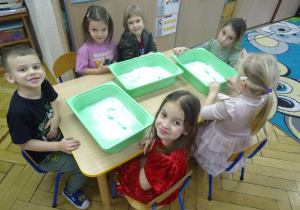 Grupa dzieci siedzy przy stoliku z plastikowymi pojemnikami.