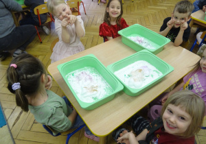 Kolejna grupa dzieci siedzy przy stoliku z plastikowymi pojemnikami.