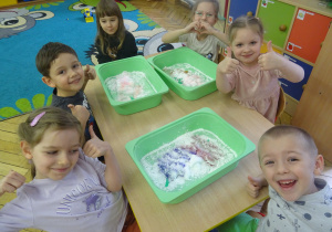 Kolejna grupa dzieci siedzy przy stoliku z plastikowymi pojemnikami.