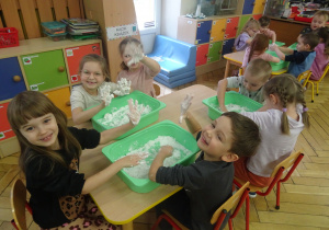 Inna grupa dzieci siedzy przy stoliku z plastikowymi pojemnikami.