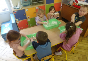 Inna grupa dzieci siedzy przy stoliku z plastikowymi pojemnikami.