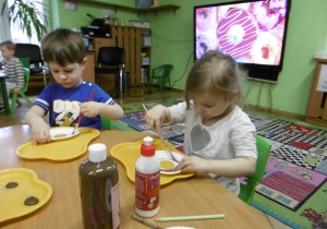 Dwoje dzieci siedzi przy stole i maluje talerzyk papierowy.