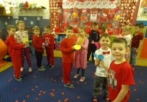 Grupa dzieci stoi ubrana na czerwona w sali przedszkolnej.