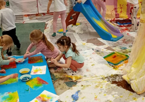 Dzieci bawią się, malują farbami i zjeżdżają na zjeżdżalni.