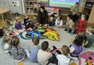 Nauczycielka siedzi na dywanie w otoczeniu dzieci. Na dywanie leżą dmuchane dinozaury.