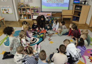 Dzieci siedzą na dywanie. Na środku znajdują się plastikowe dinozaury.