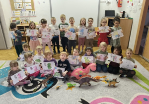 Grupa dzieci stoi razem obok siebie i trzyma w rękach wykonane prace plastyczne.