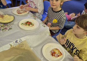 Grupa dzieci siedzi przy stole z przygotowanymi przez siebie pizzami.