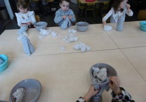 Dzieci siedzą przy stole i wypychają watą szare skarpetki.