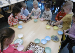 Grupa dzieci siedzi wokół stołu i przebiera w kolorowych guzikach.