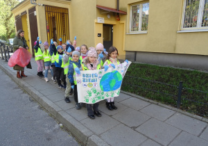 Grupa dzieci stoi przed wejściem do przedszkola i trzyma wykonany przez siebie plakat.