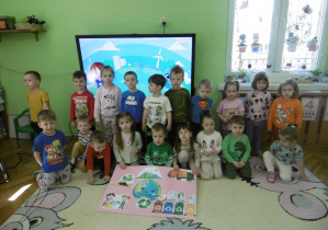 Grupa dzieci siedzi obok siebie. Przed nimi plakat