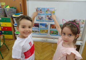 Dwoje dzieci stoi obok siebie i ręką wskazuje na ilustrację.