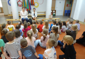 Dzieci siedzą na dywanie przed osoboiami z Sanepidu.