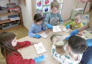 Dzieci siedzą przy stoliku i kroją warzywa.