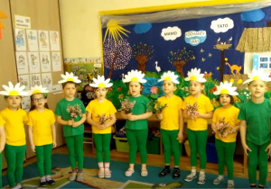 Żaczki ustawione w półkolu w zielonych geterkach i żółtych bluzeczkach podczas występów z okazji Dnia mamy i Taty