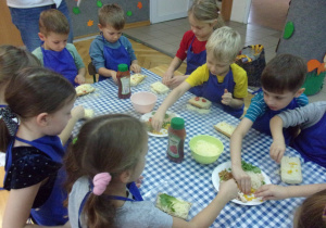 Juniorzy w fartuszkach przy stolikach przygotowują zapiekanki